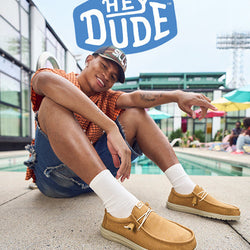 A young man sitting poolside in a relaxed pose, wearing tan Hey Dude shoes, white socks, denim shorts, and a cap, smiling and pointing playfully, with the Hey Dude logo above.