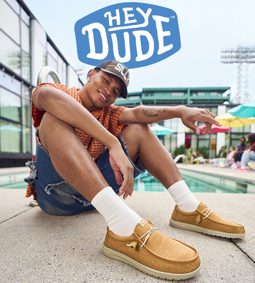 A young man sitting poolside in a relaxed pose, wearing tan Hey Dude shoes, white socks, denim shorts, and a cap, smiling and pointing playfully, with the Hey Dude logo above.