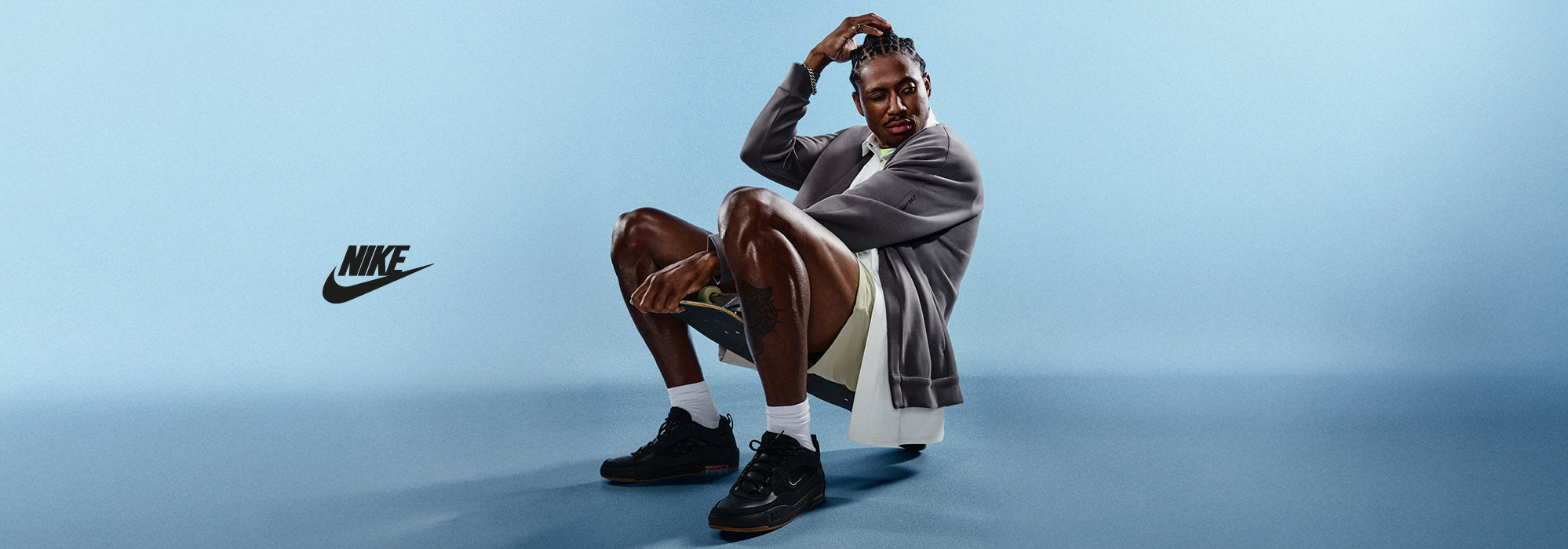 A stylish man in a relaxed seated pose wearing Nike sneakers and streetwear, set against a clean blue background with the Nike logo displayed prominently.