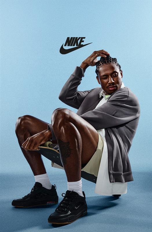 Portrait shot of a man dressed in Nike apparel and sneakers, sitting confidently with a skateboard, framed by a minimal blue backdrop and Nike logo.