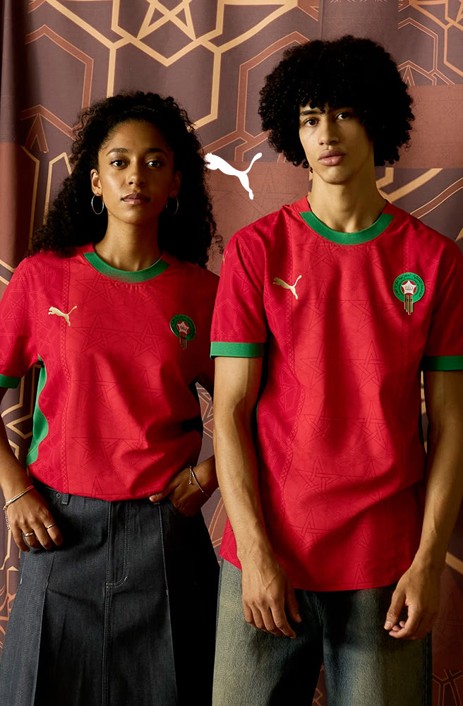 A woman and man wearing the official red Puma jerseys of the Moroccan national football team, standing in front of a traditional Moroccan patterned background.